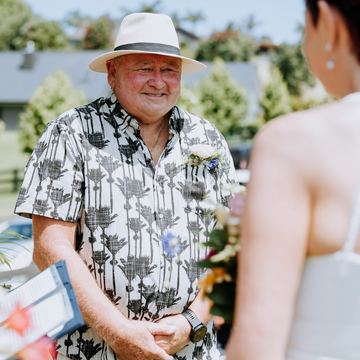 The groom in a sunhat and patterned shirt smiles at the bride