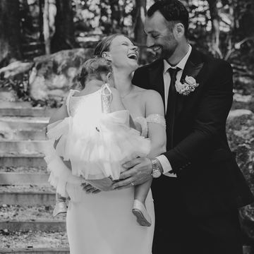Black-and-white photo of a bride laughing as she holds a little girl, with the groom beside her