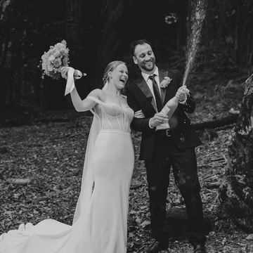 Black-and-white photo of a bride and groom celebrating with champagne in a forest clearing