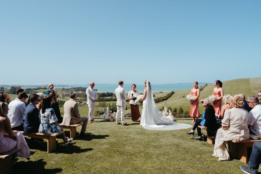 Guests seated on benches watch a hilltop wedding ceremony with the sea behind