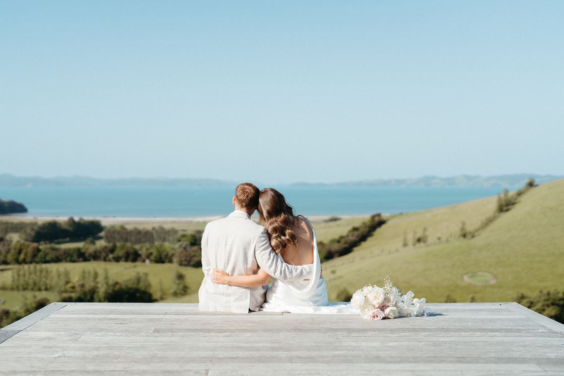 A newly married couple sit side by side on a deck looking out over hills to the sea